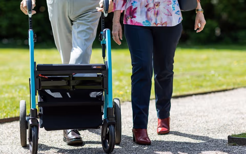Two seniors walk outdoors, using a mobility walker along a sunny path.