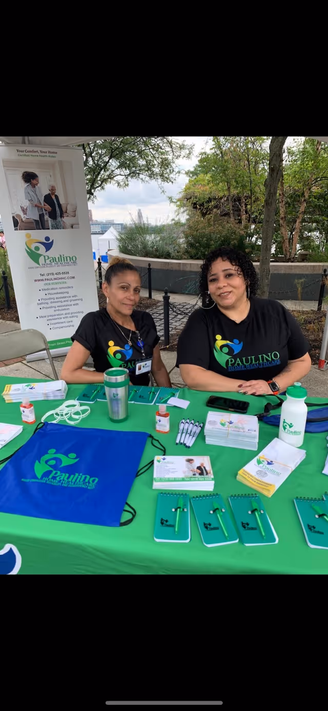 Two women sit at a green outdoor booth with a Paulino Home Healthcare banner.