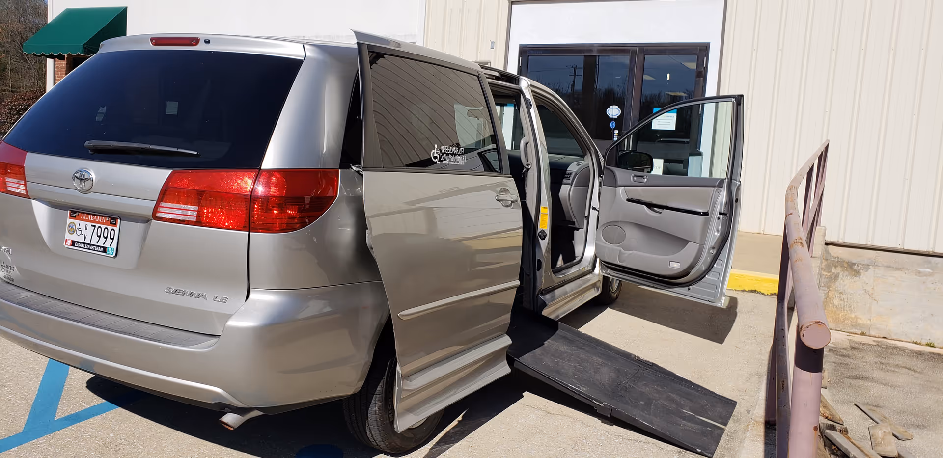 Silver wheelchair-accessible van with ramp extended parked by a facility entrance.