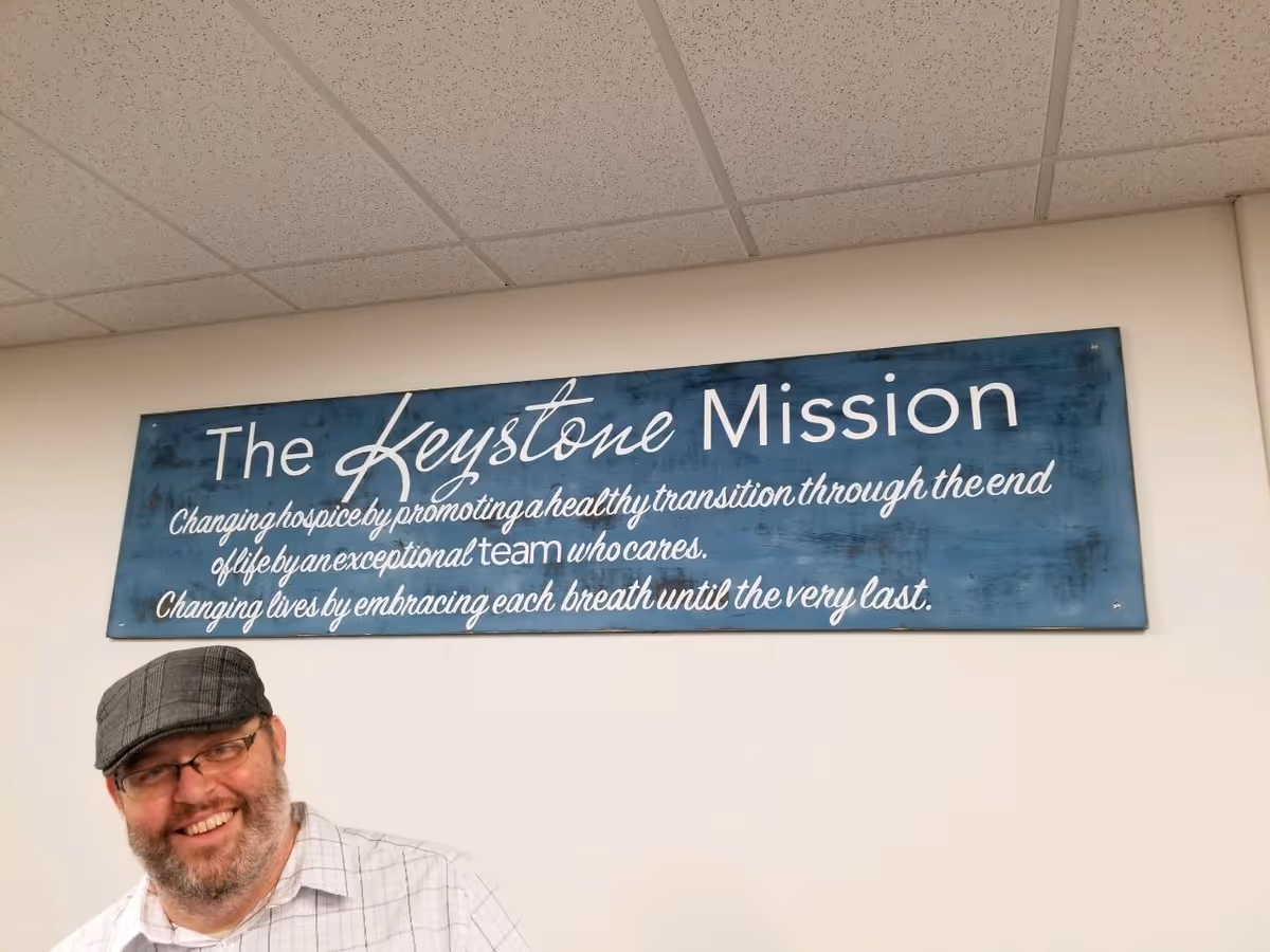 Smiling man wearing a cap stands beside a blue sign reading The Keystone Mission.