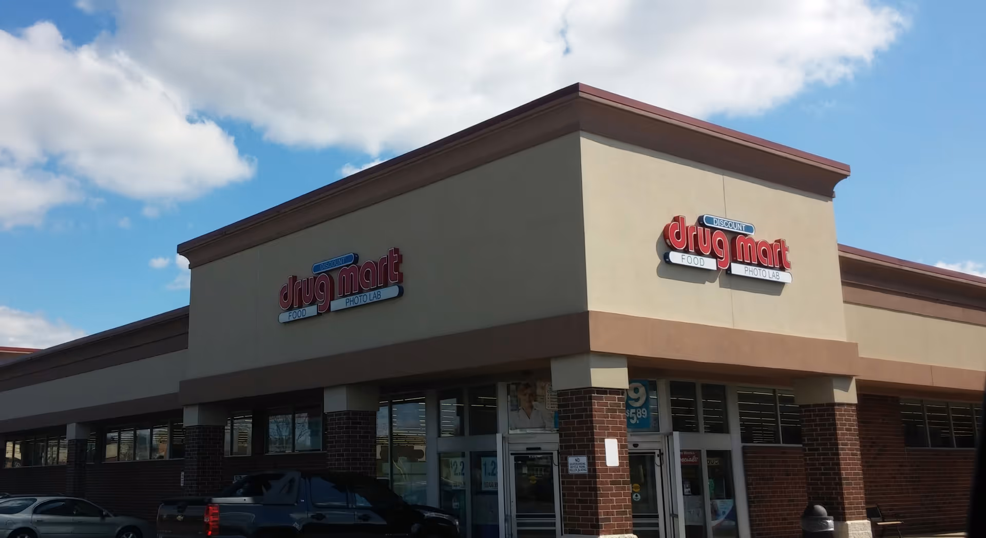 Exterior view of a Discount Drug Mart storefront with brick accents and parked cars.