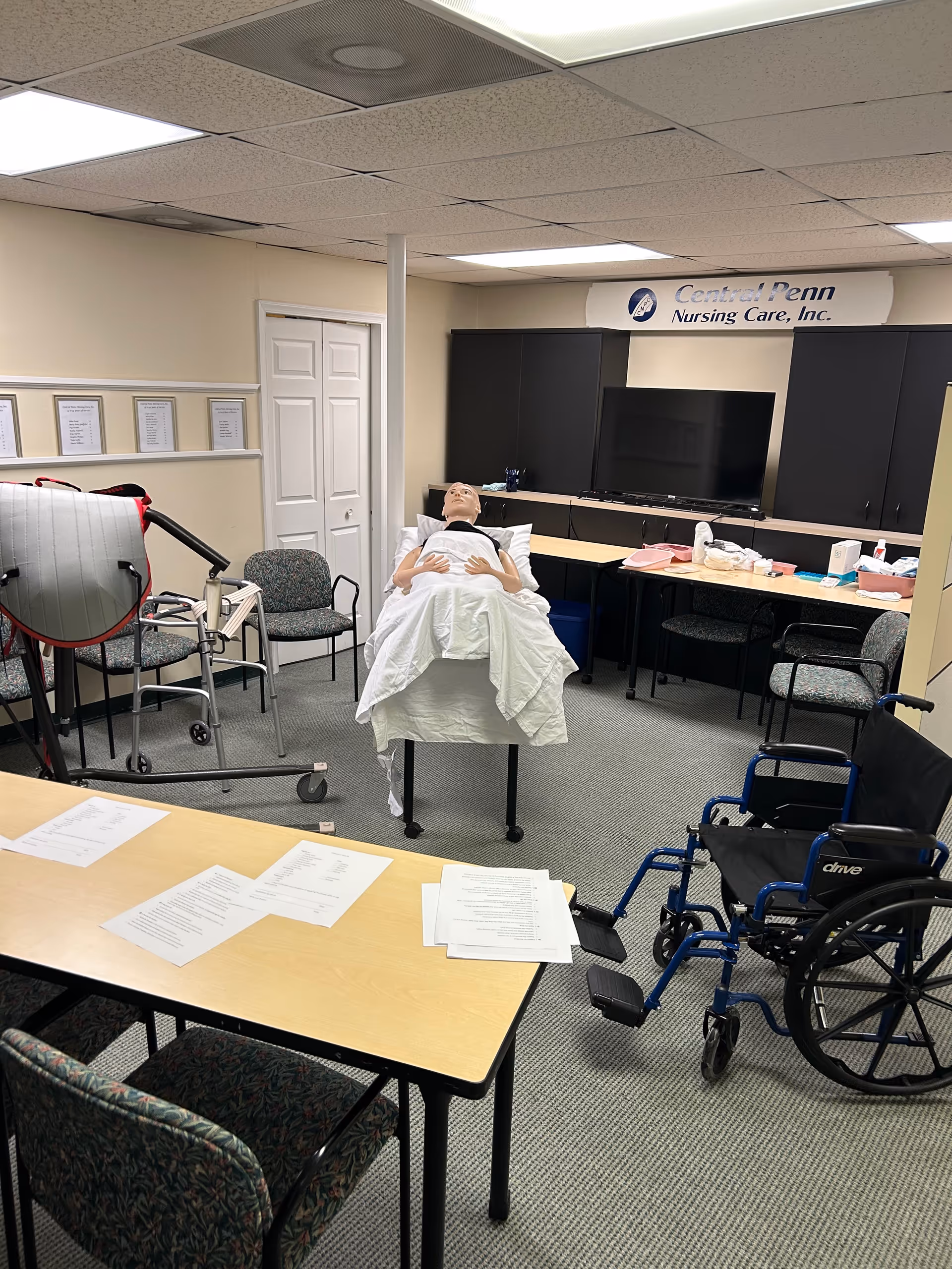 Medical training setup in a nursing care room with a mannequin on a bed and chairs.