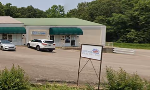 Exterior view of At Home Healthcare facility with parked cars and a sign in the foreground.