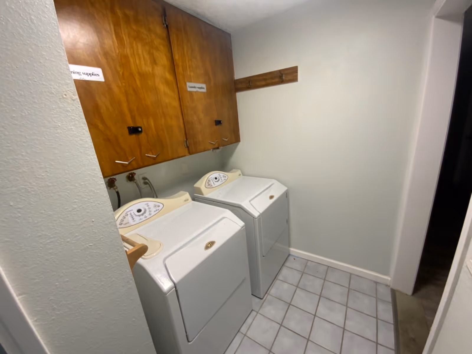 Laundry room with two top-loading washers, wooden cabinets, and tiled floor.