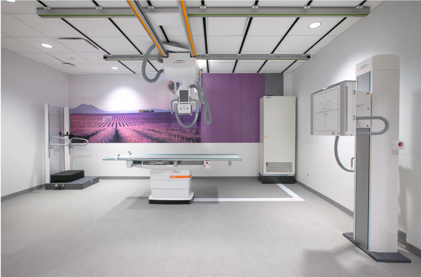 Empty radiology suite with X-ray table, ceiling-mounted imaging equipment, and purple mural.