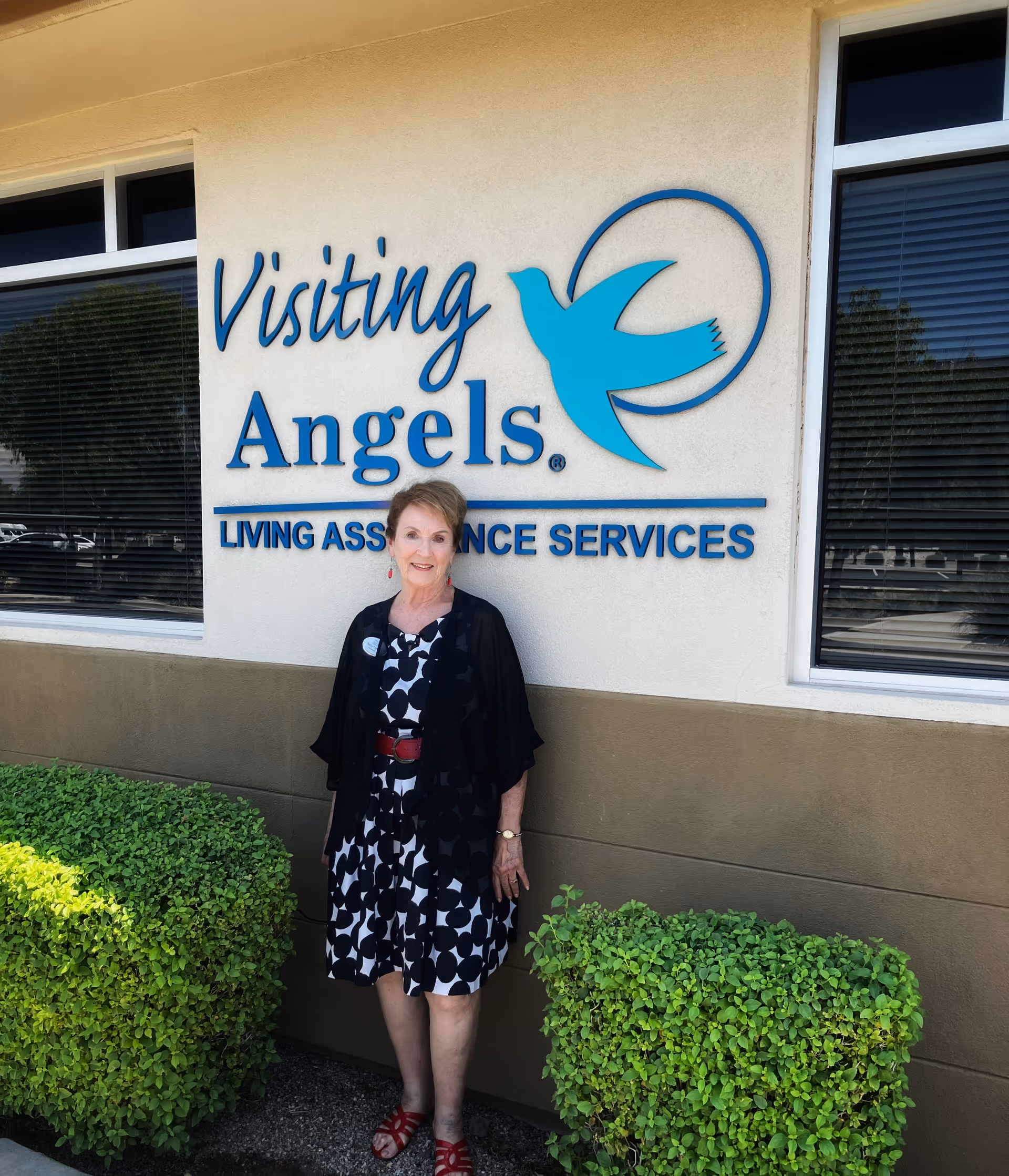 Smiling woman stands outside Visiting Angels sign at a senior home care facility.