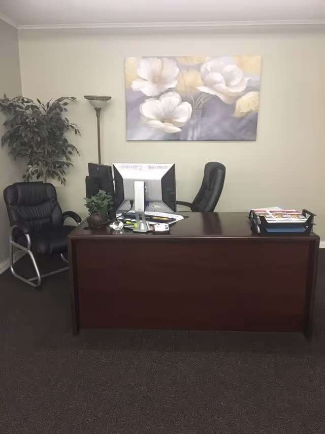 Office reception area with desk, two computer monitors, chairs, plant, and wall art.