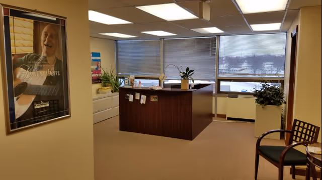 Reception desk in a senior living facility lobby with plants and large windows.
