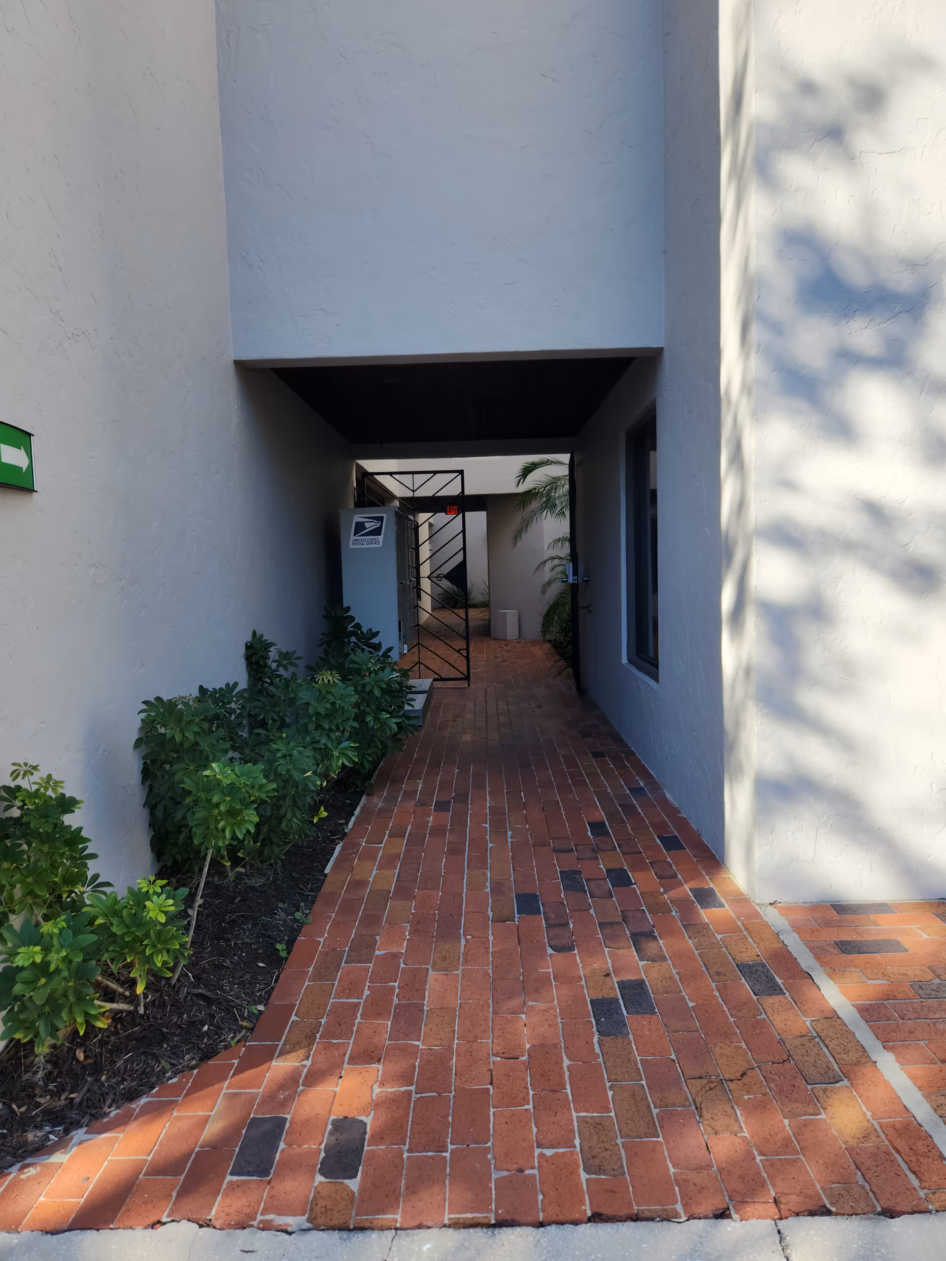 Brick-paved outdoor walkway between light gray walls with shrubs on the left.