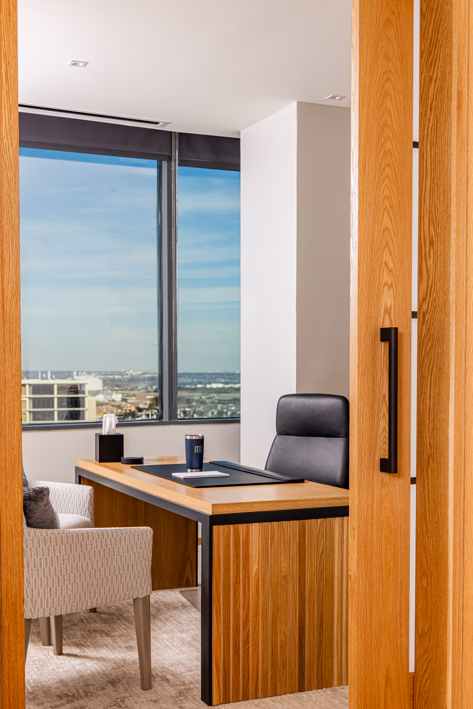 Interior office area with a wooden desk, black leather chair, and large window view.
