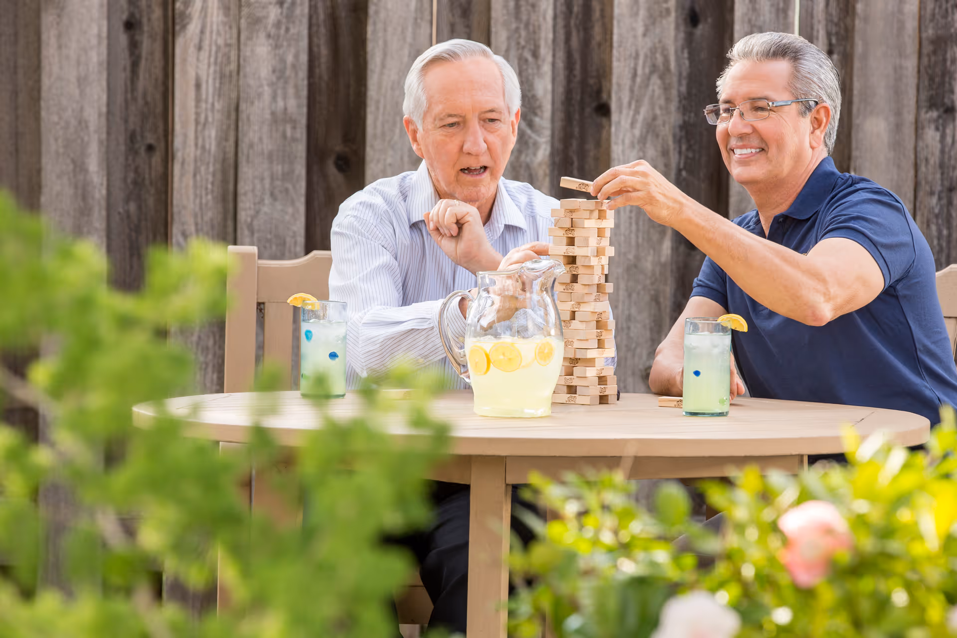 Two elderly men enjoy a game of Jenga outdoors at a table with lemonade.