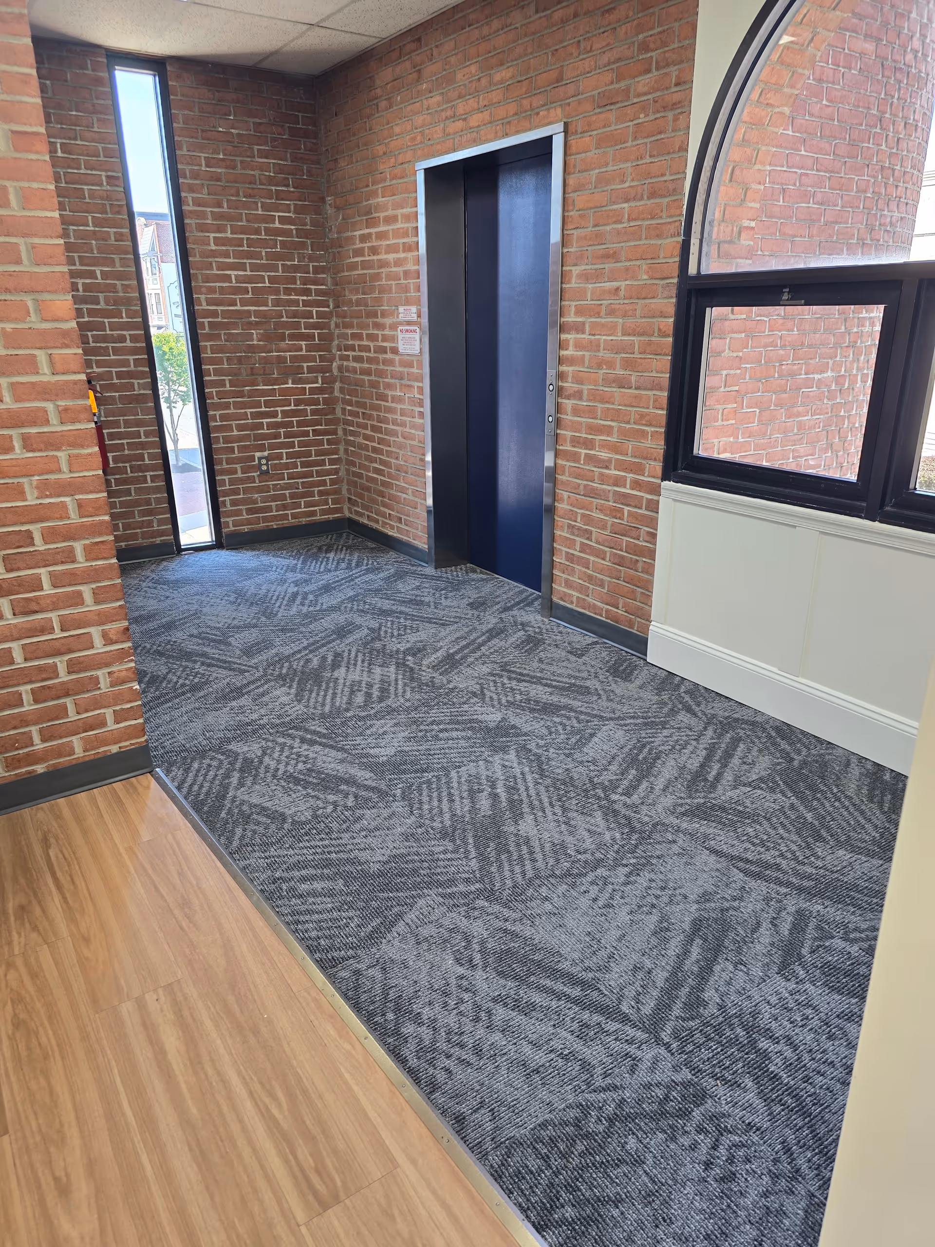 Interior hallway with brick walls, a blue elevator, arched window, and patterned carpet.