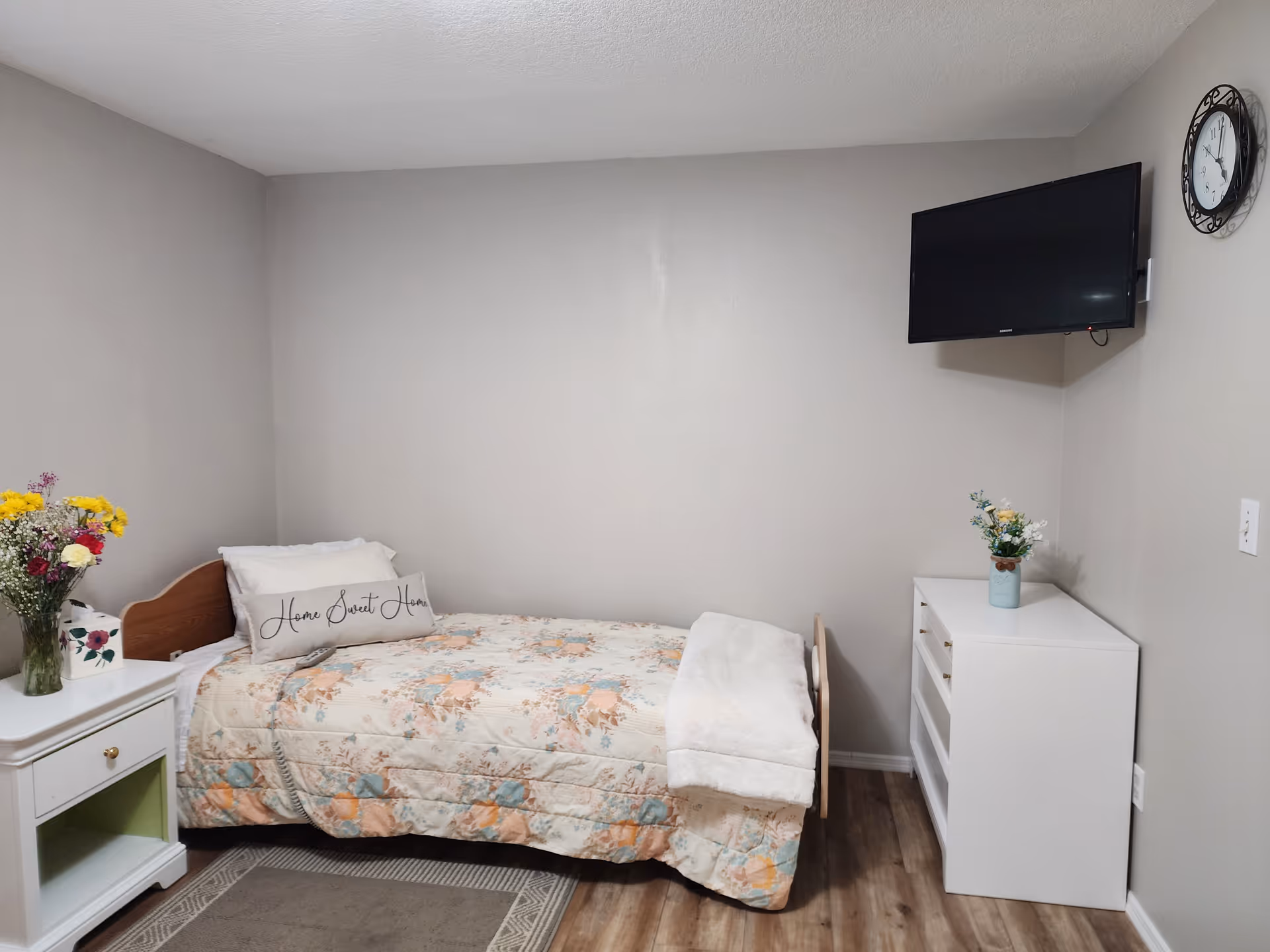 Cozy single bedroom with bed, dresser, mounted TV, clock, and fresh flower decor.