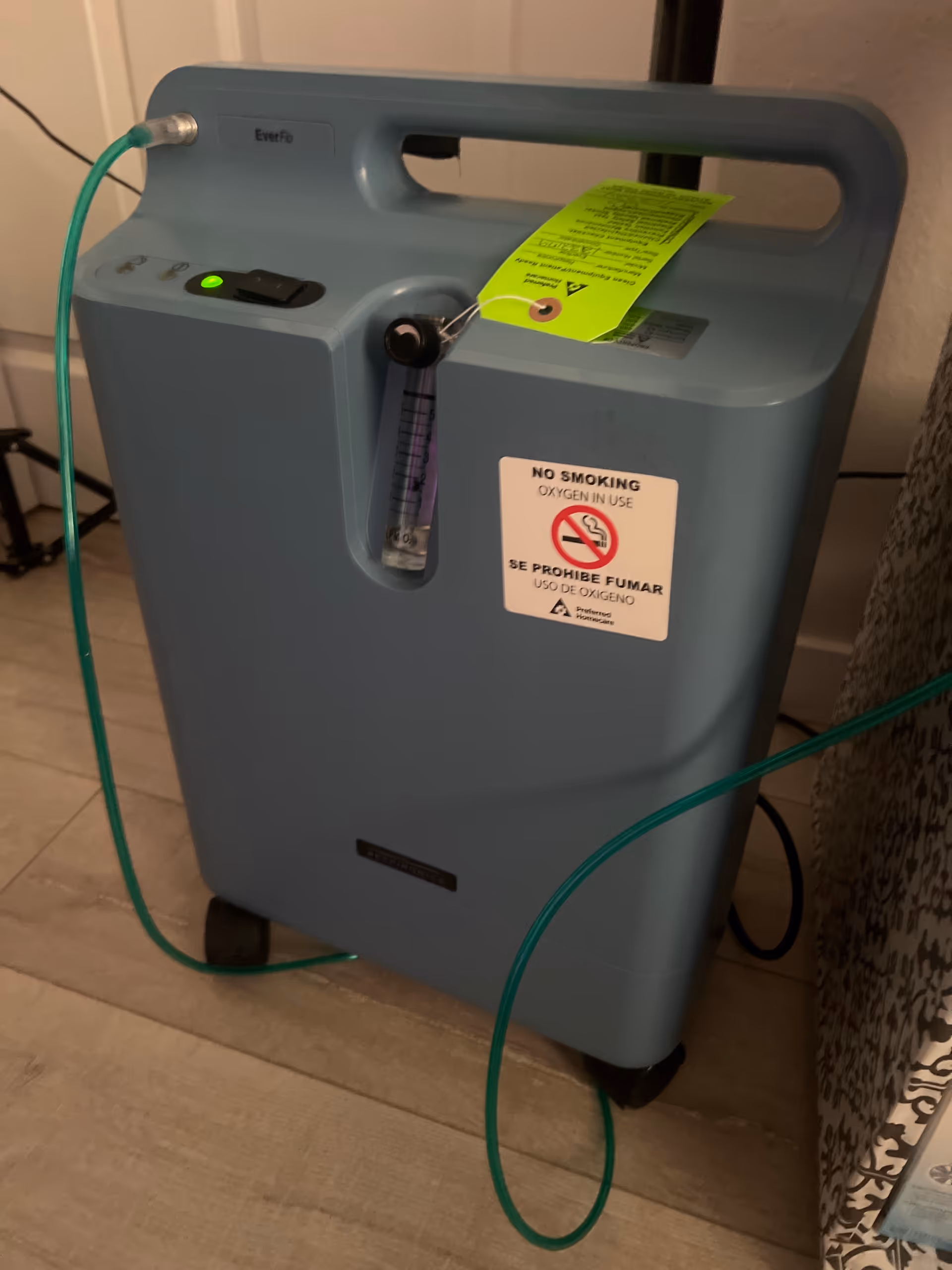 Blue portable oxygen concentrator on wheels with green tubing indoors.