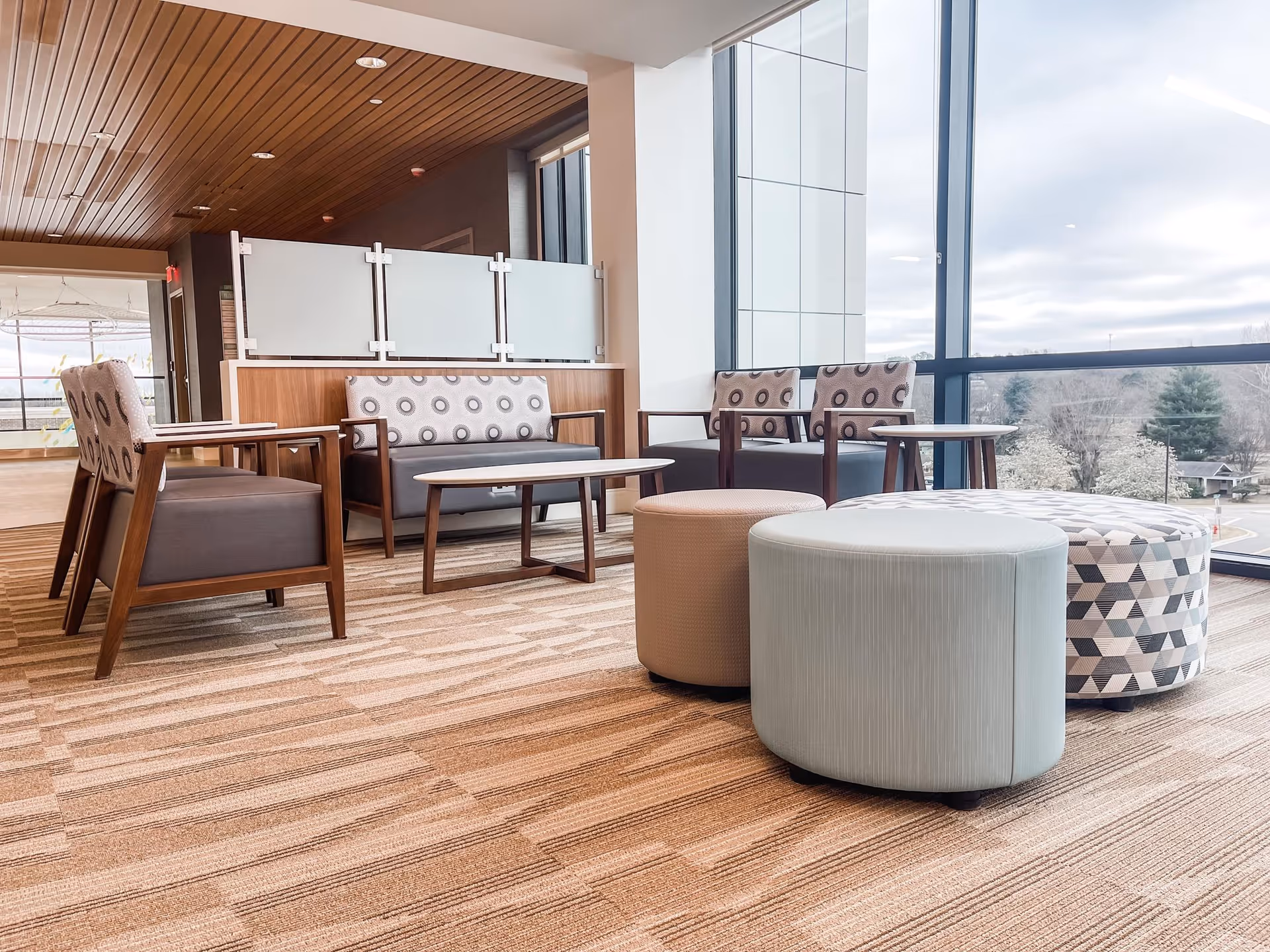Modern senior living lounge with wooden chairs, patterned ottomans, and large glass windows.