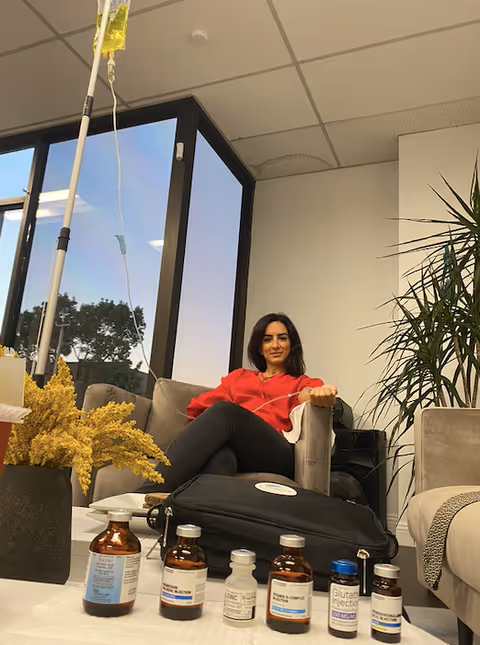 A woman in red sits on a sofa in a lounge with an IV pole nearby.