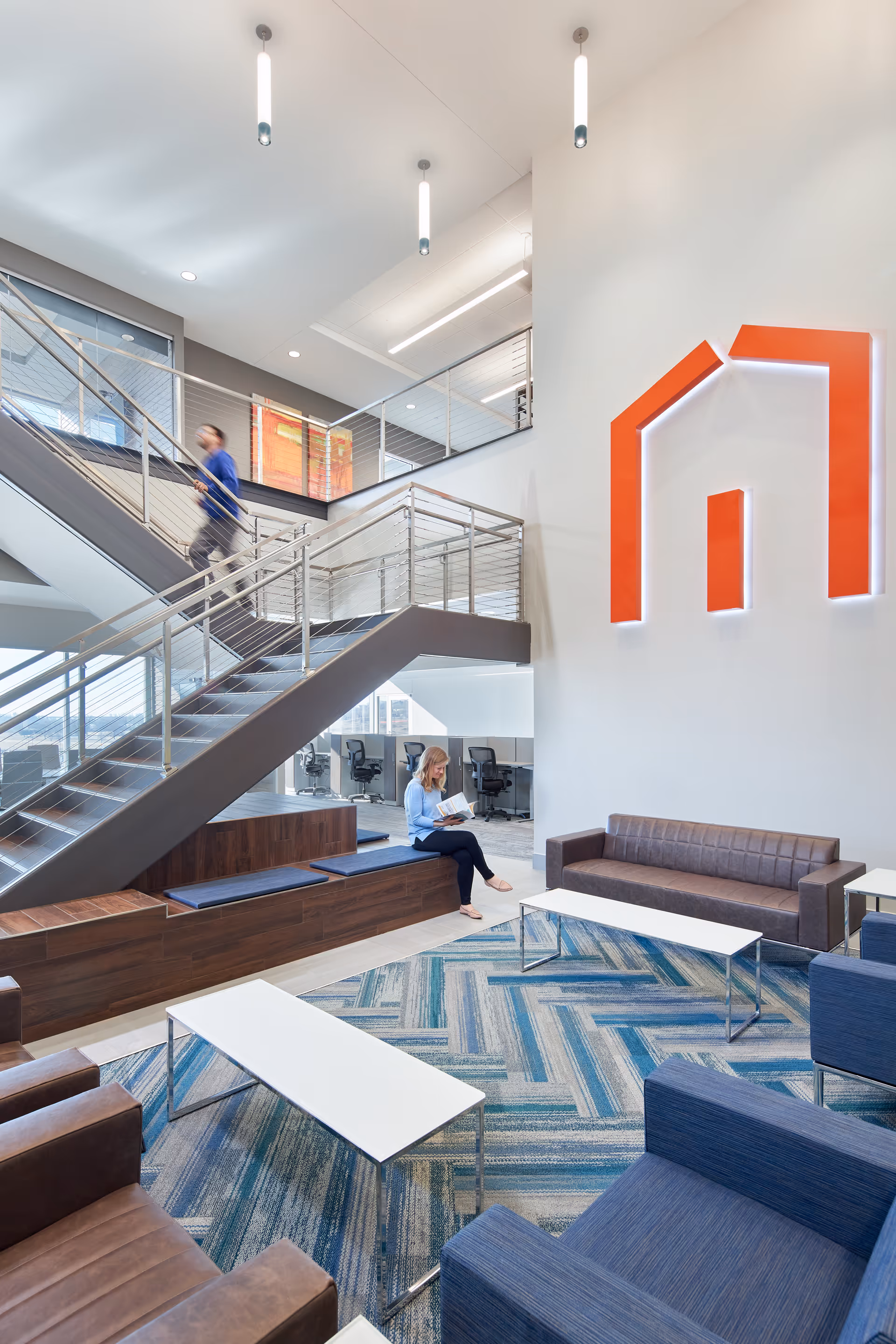 Modern corporate lobby with blue patterned carpet, seating, stairway, and orange logo on wall.