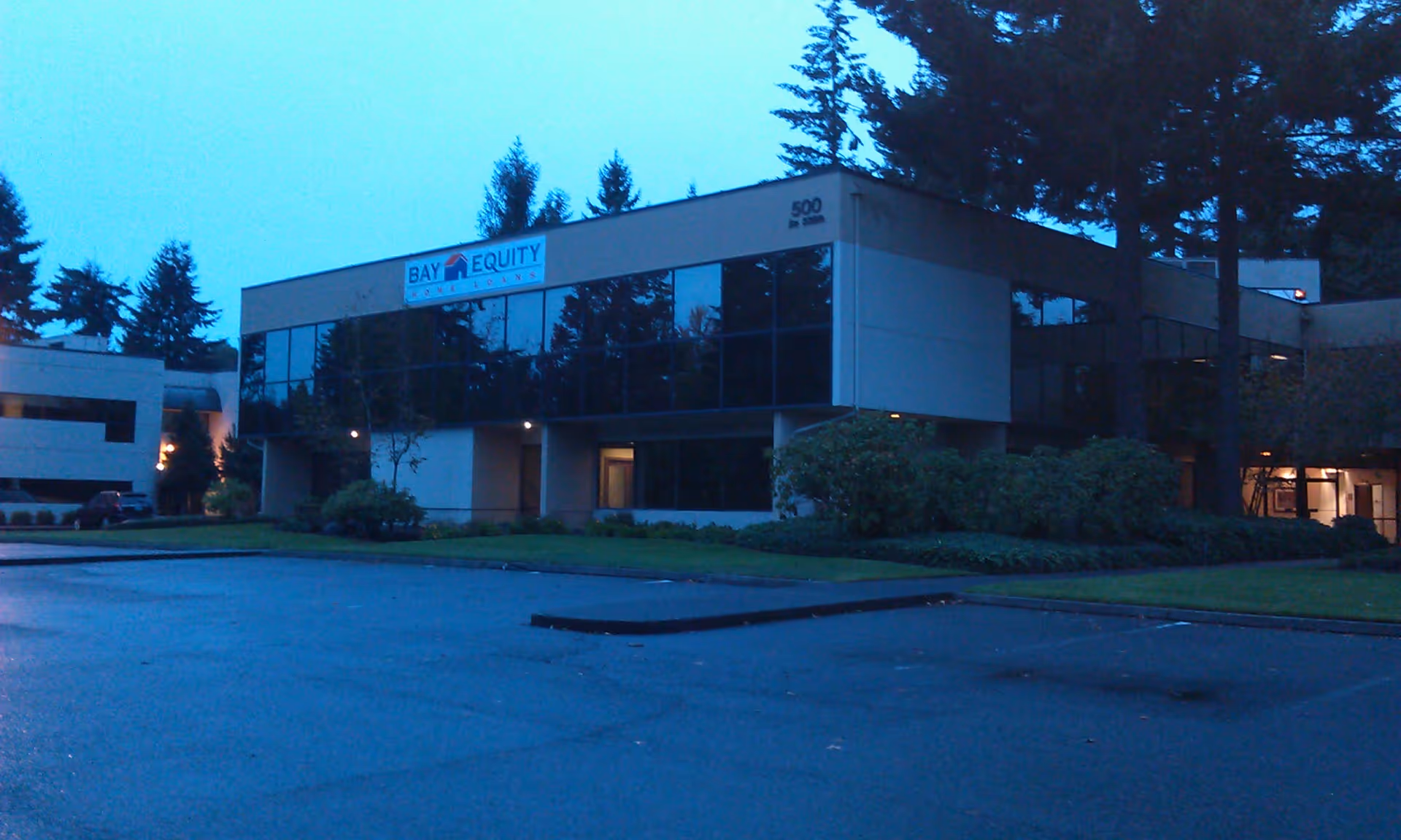 Exterior view of a modern, glass-front building with trees and a parking lot.
