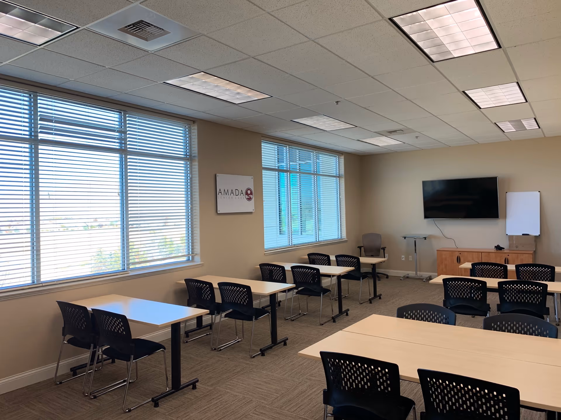 Bright classroom-style meeting room with tables, chairs, windows, and a wall-mounted TV.