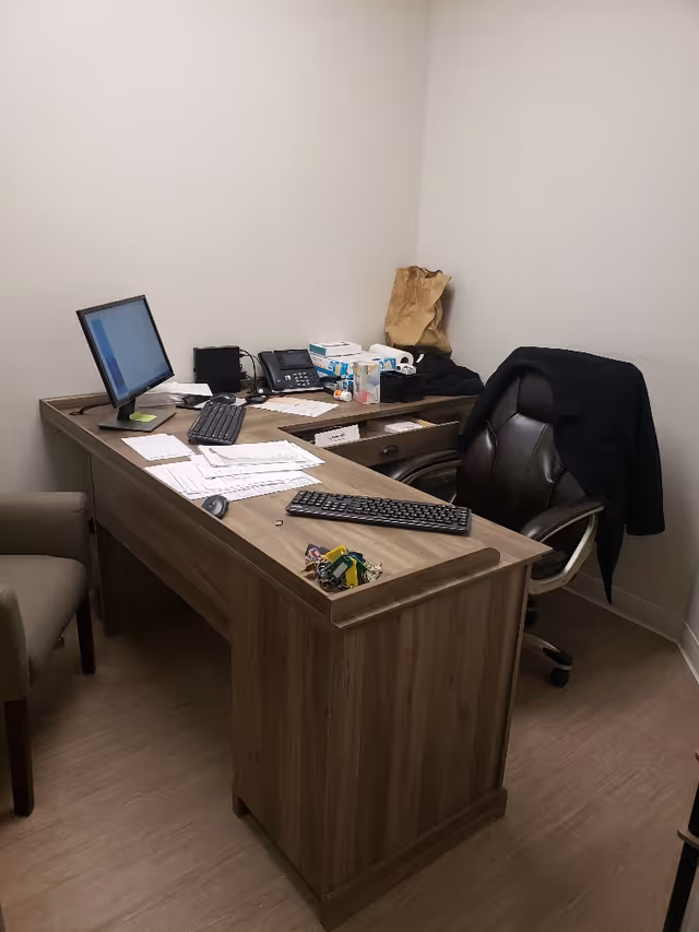 A small office setup with a wooden desk, computer, keyboard, papers, and a chair.
