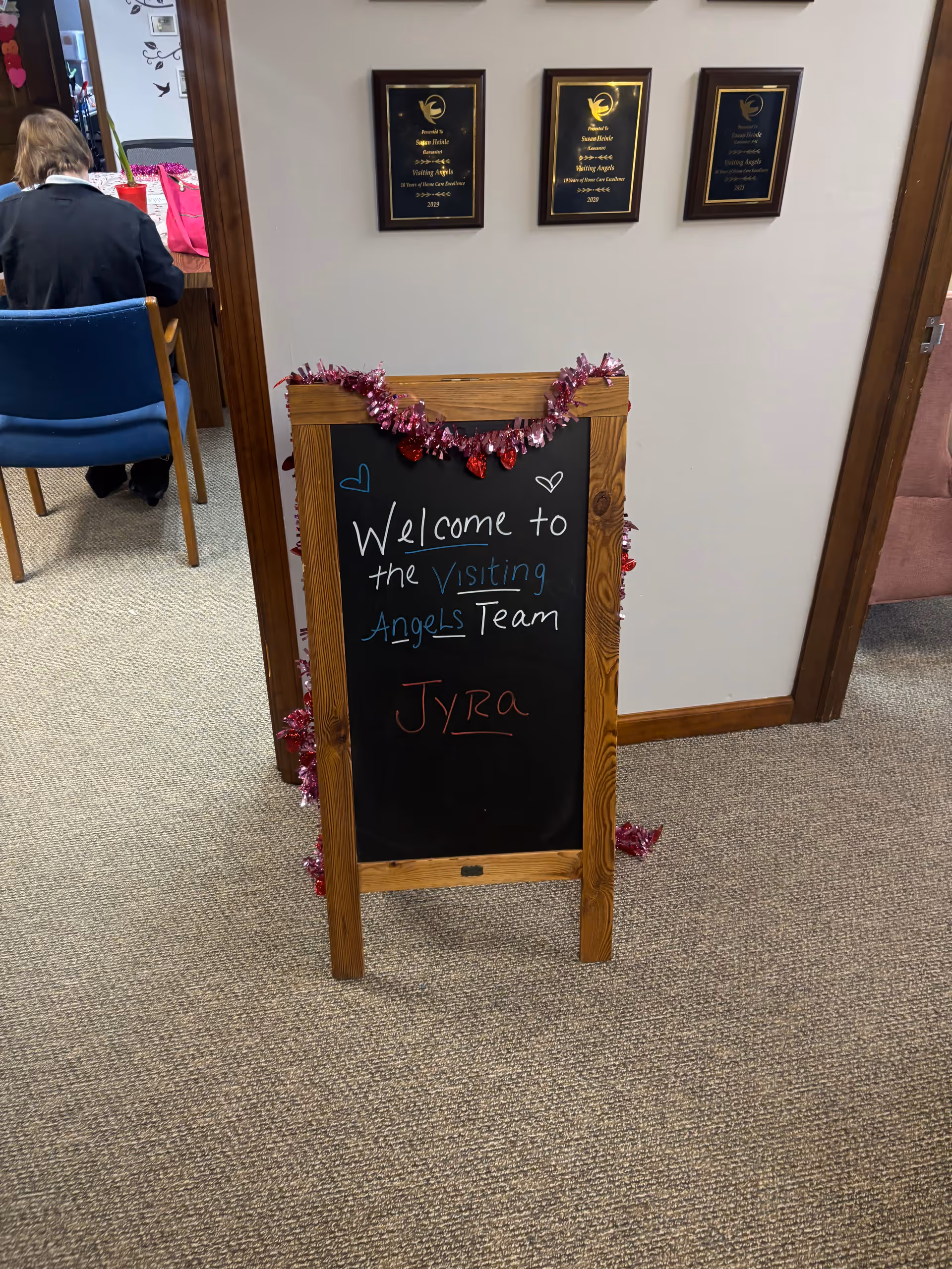 Indoor hallway with chalkboard welcome sign for Visiting Angels Team and a seated staff member.