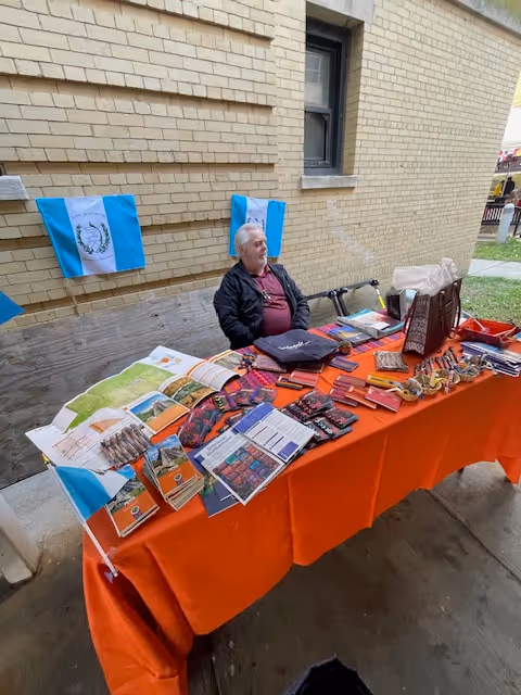 A man sits behind an orange table of brochures outdoors beside a brick building.