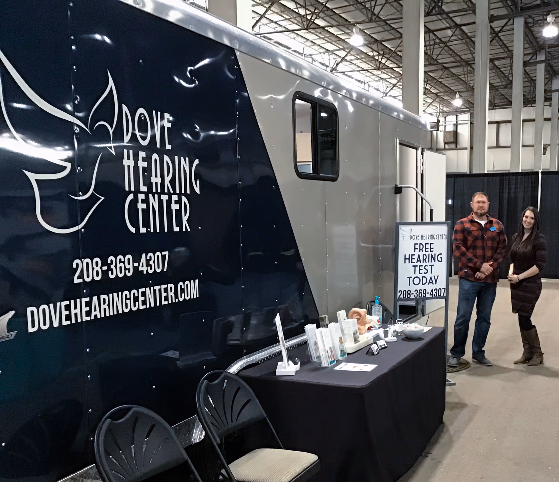 Dove Hearing Center trailer booth inside a large hall with two staff near a display table.