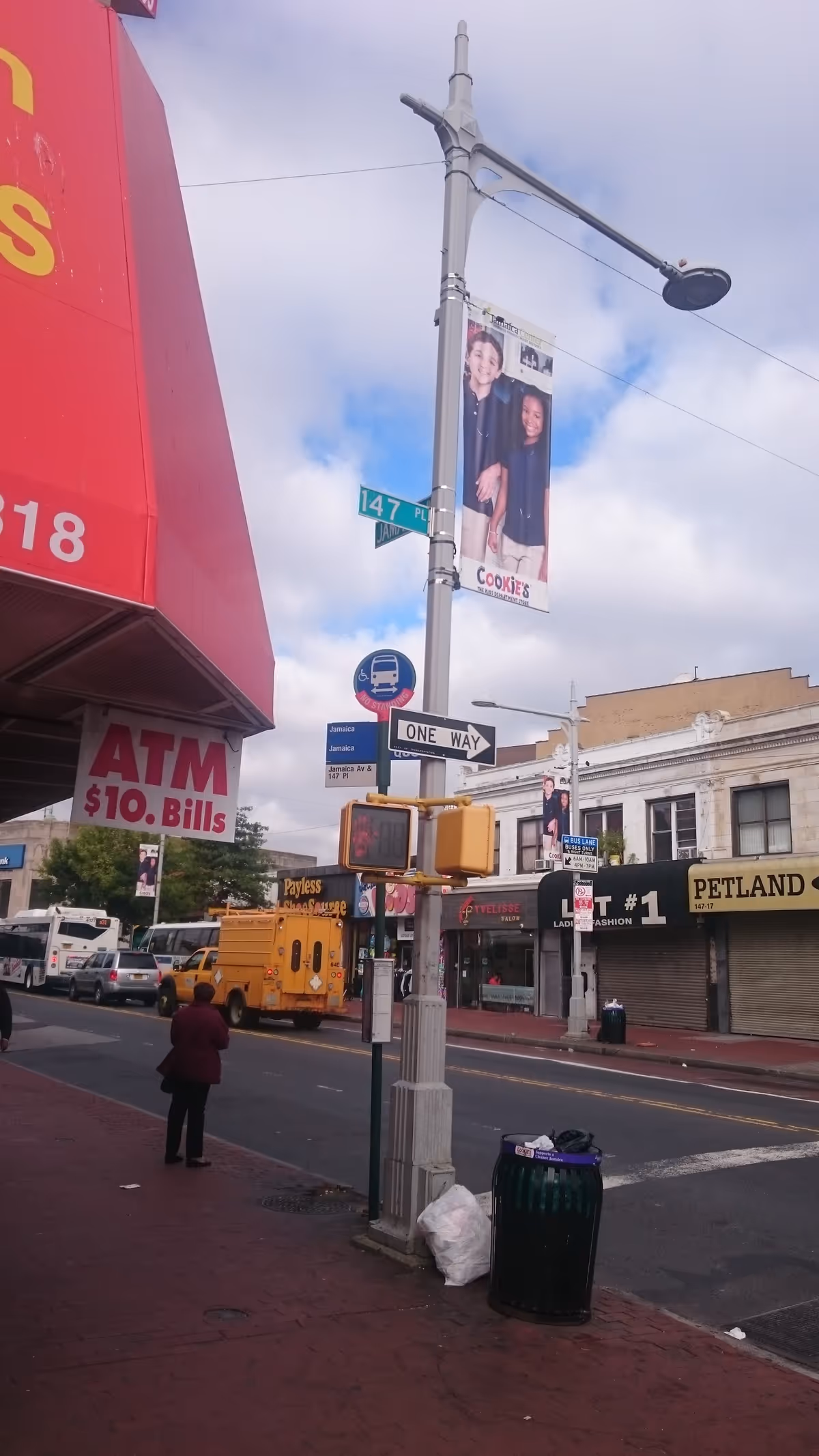 Urban street corner with storefronts, street signs, a lamppost banner, and a pedestrian.