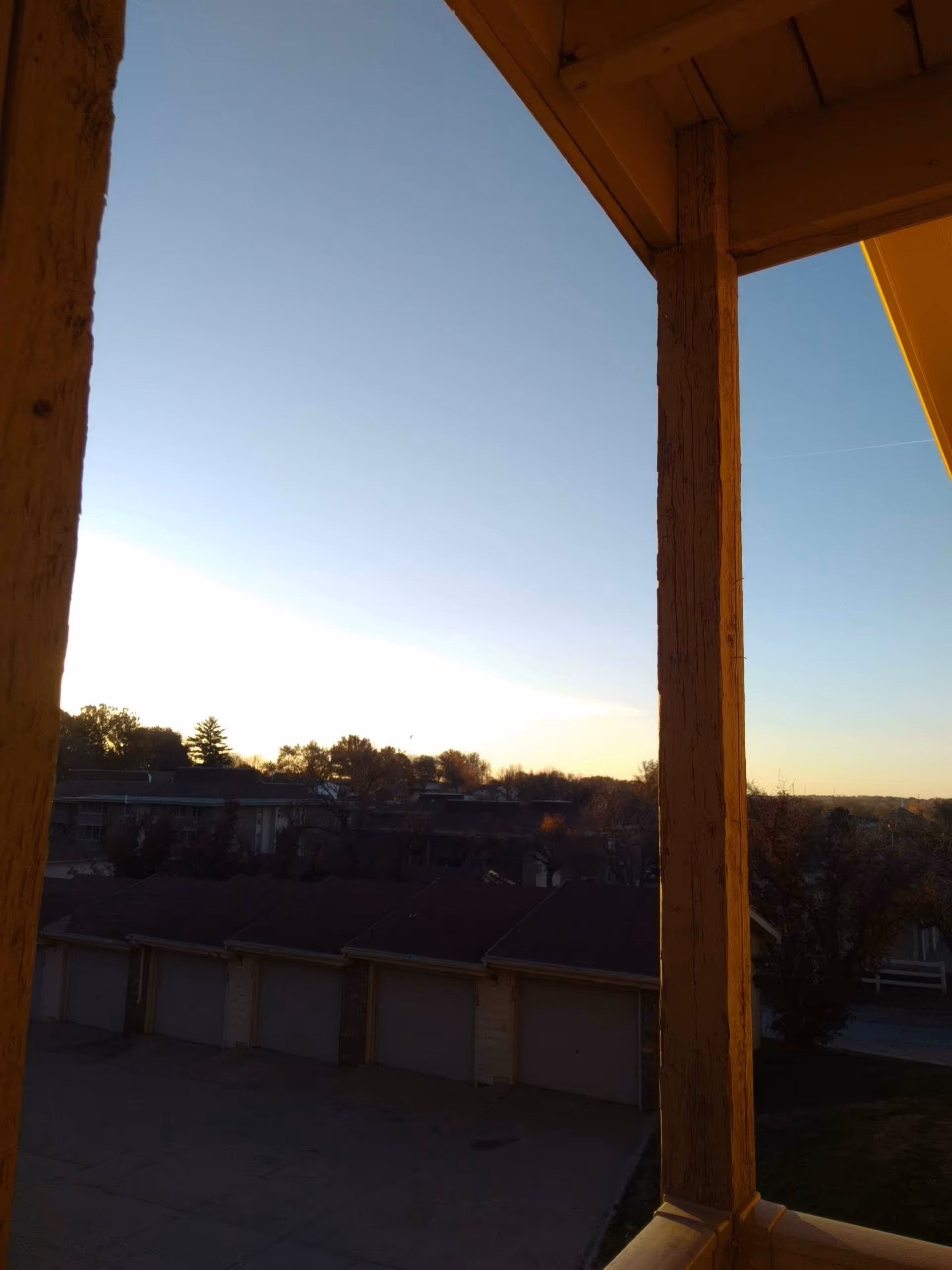 Sunlit exterior balcony with wooden posts overlooking row of garages and distant trees.