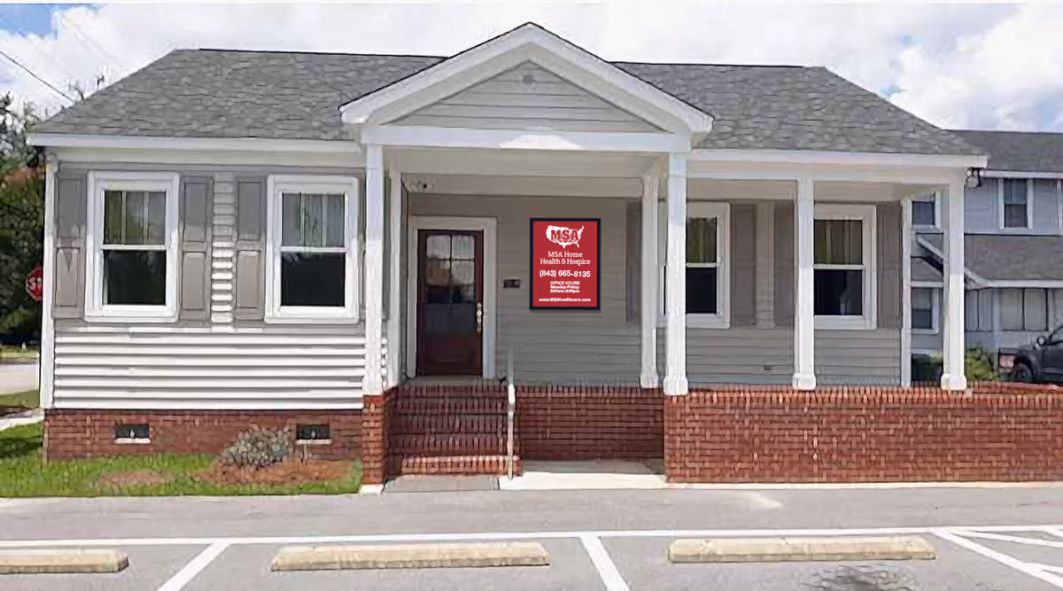 Single-story light gray facility with white columns and a red brick foundation, front entrance, sign.