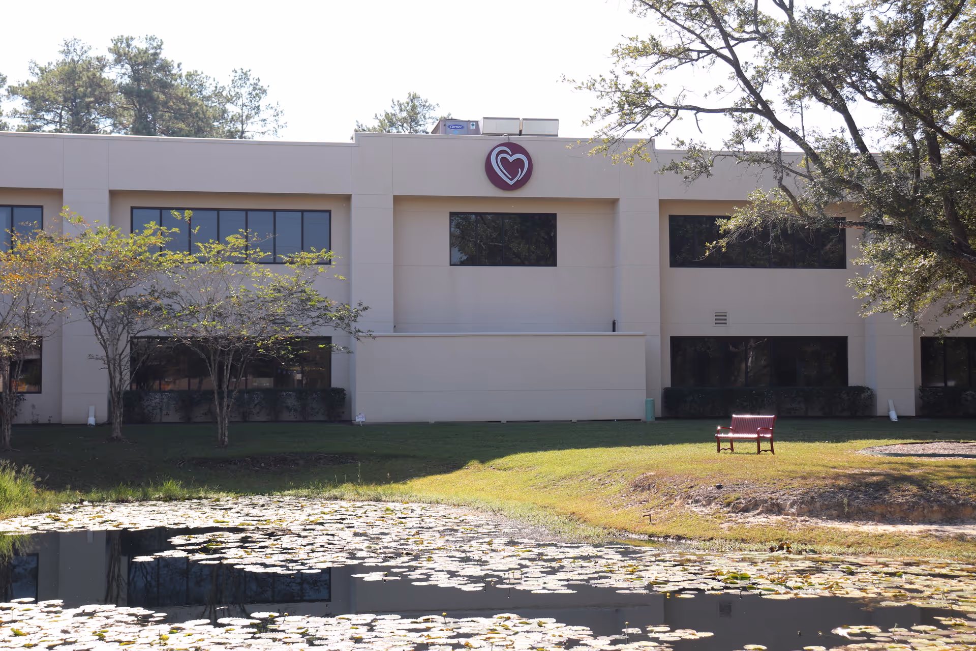 Two-story beige facility with heart logo, trees, lawn, and a pond with a bench.