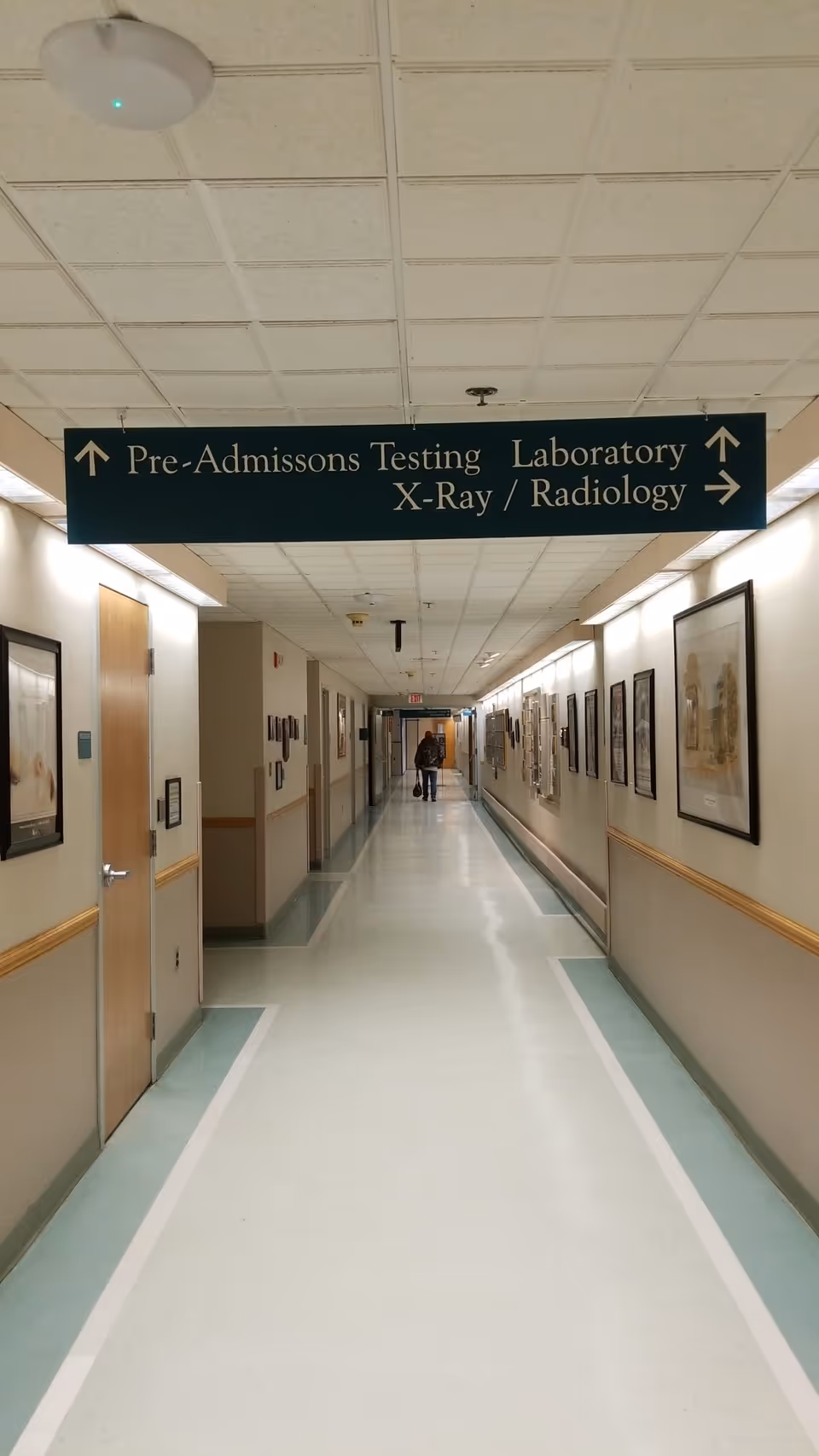 Long hospital corridor with framed wall art and an overhead directional sign.