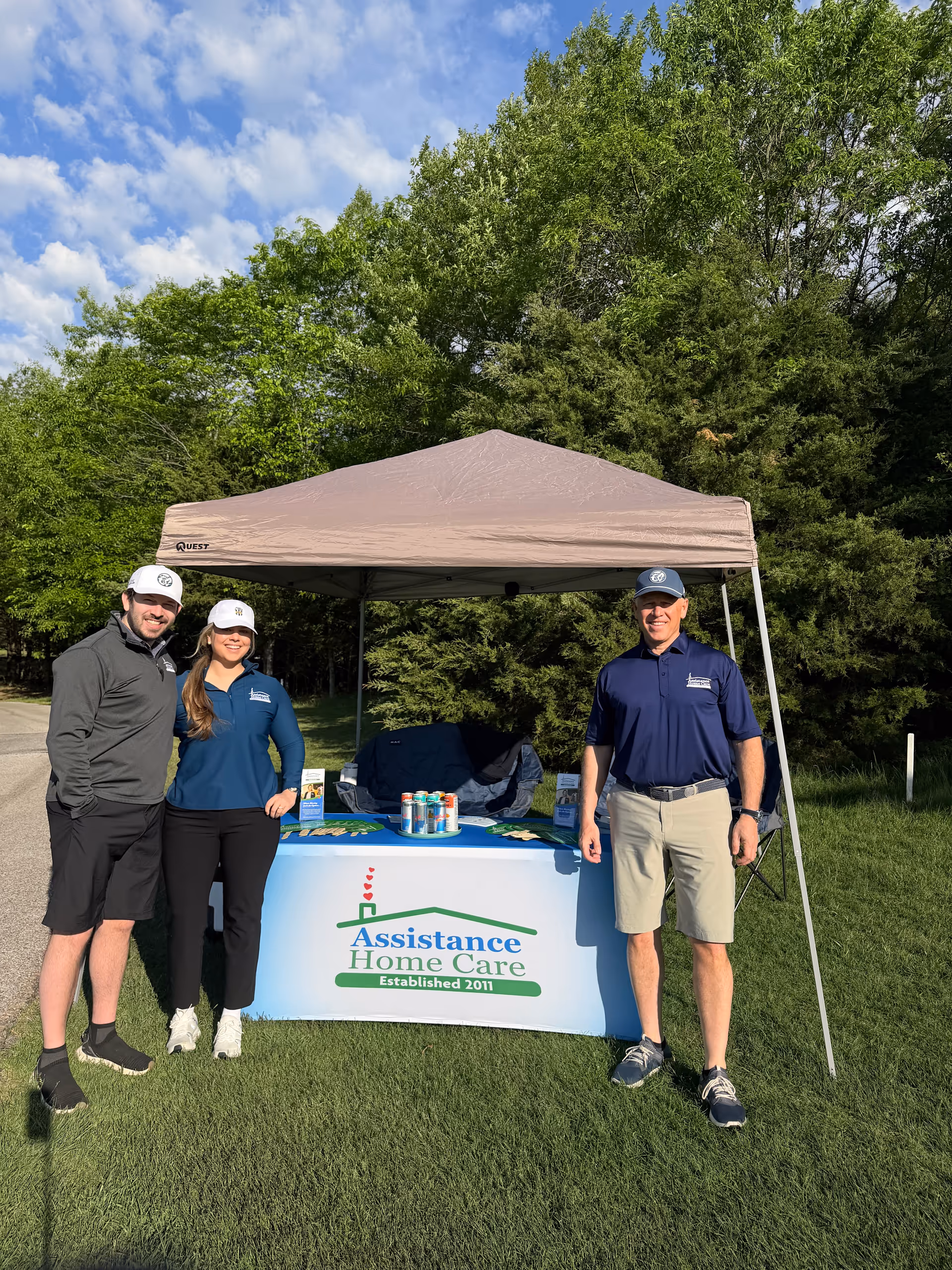 Three people stand beside a tented promotional table with Assistance Home Care signage outdoors.