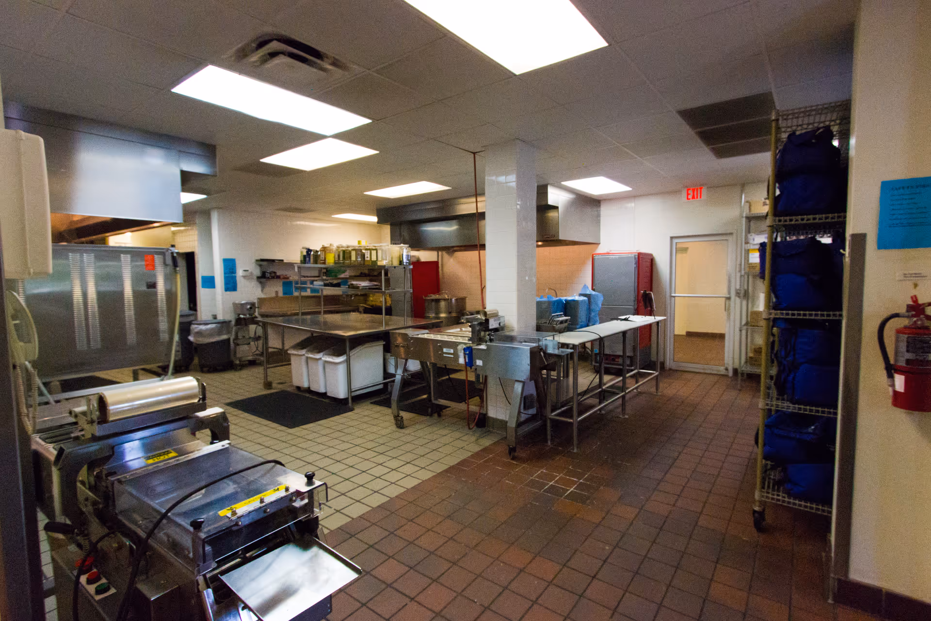 Commercial kitchen with stainless steel appliances, prep tables, and shelving in a tiled facility.