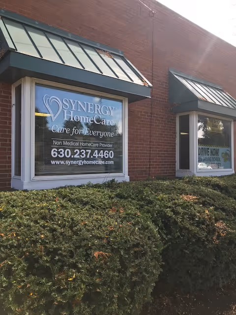 Brick storefront with Synergy HomeCare sign in windows and trimmed hedge in front.