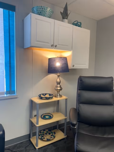Corner of a living room with a lamp, cabinets, shelves of dishes, and a dark recliner.
