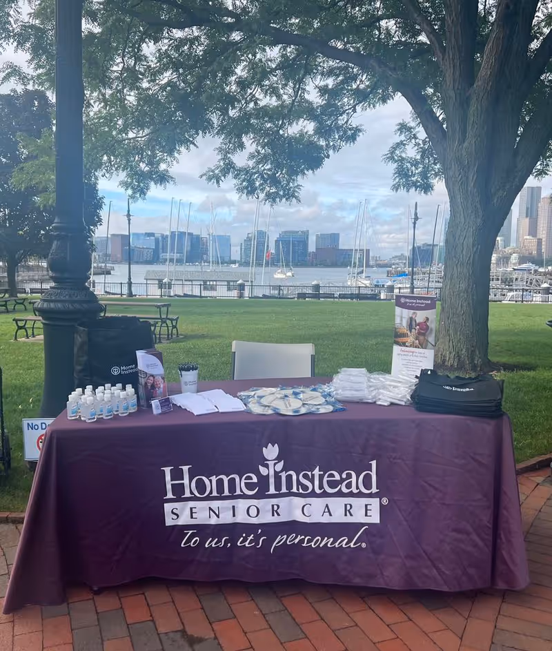 Outdoor park table with purple Home Instead tablecloth, brochures, and sanitizer near waterfront.