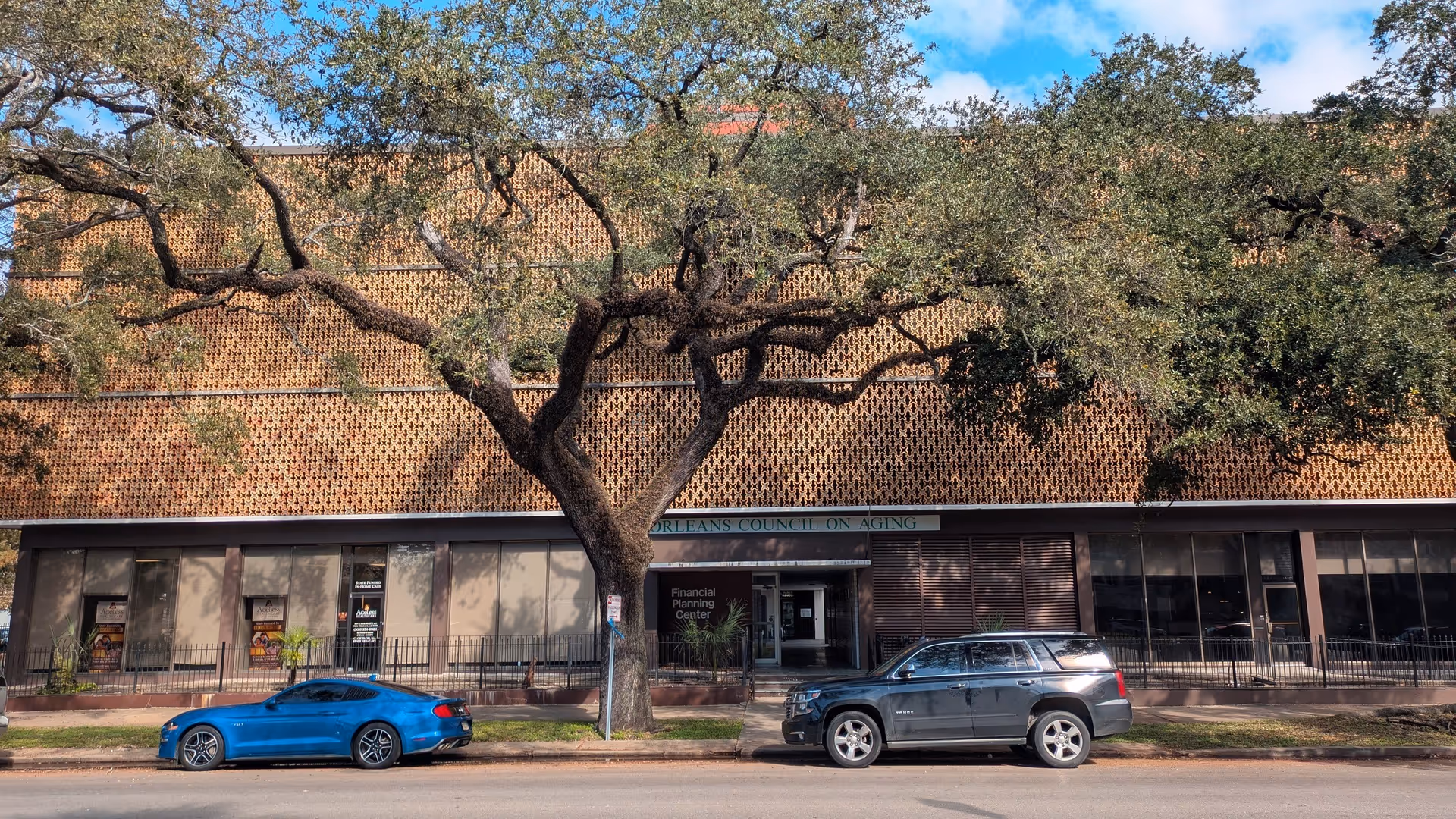 Exterior of a brick building with large windows, a tree, and parked cars.