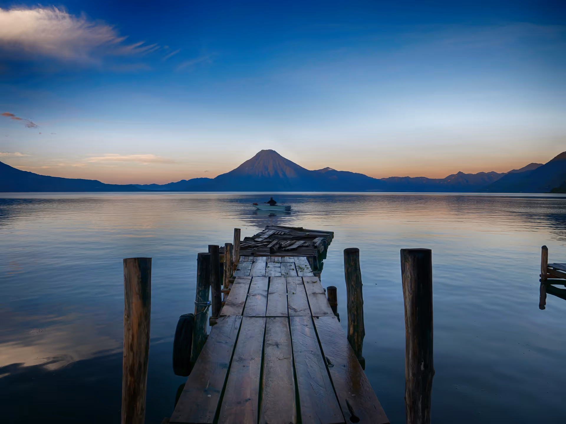 Weathered wooden dock extends over a calm lake toward distant mountains at sunset.