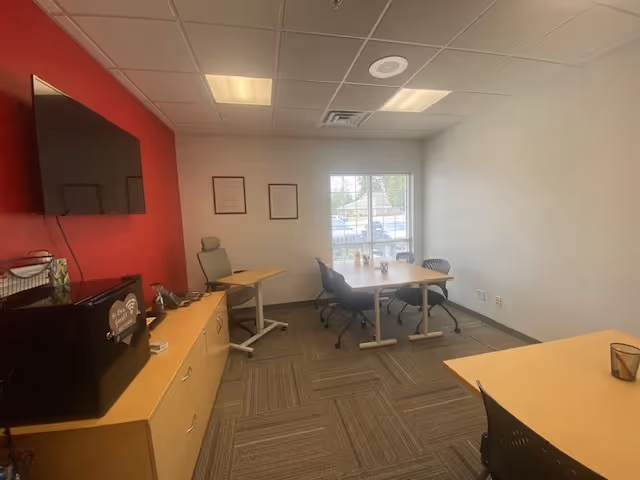 A small interior meeting area with a rectangular table, chairs, red accent wall, and coffee station.