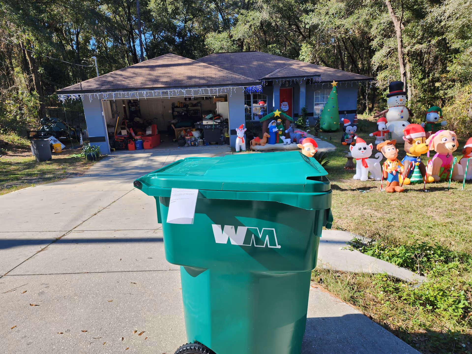Front view of a house with Christmas inflatables, decorations, and a green trash bin in driveway.