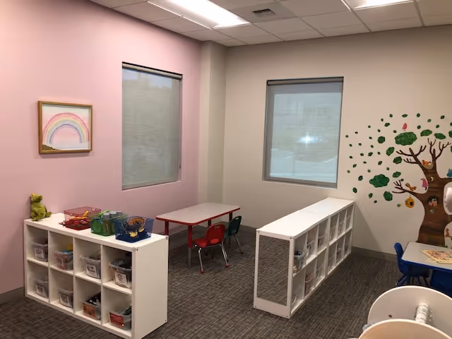 Bright interior activity room with cubbies, kid sized tables, and a tree mural on the wall.