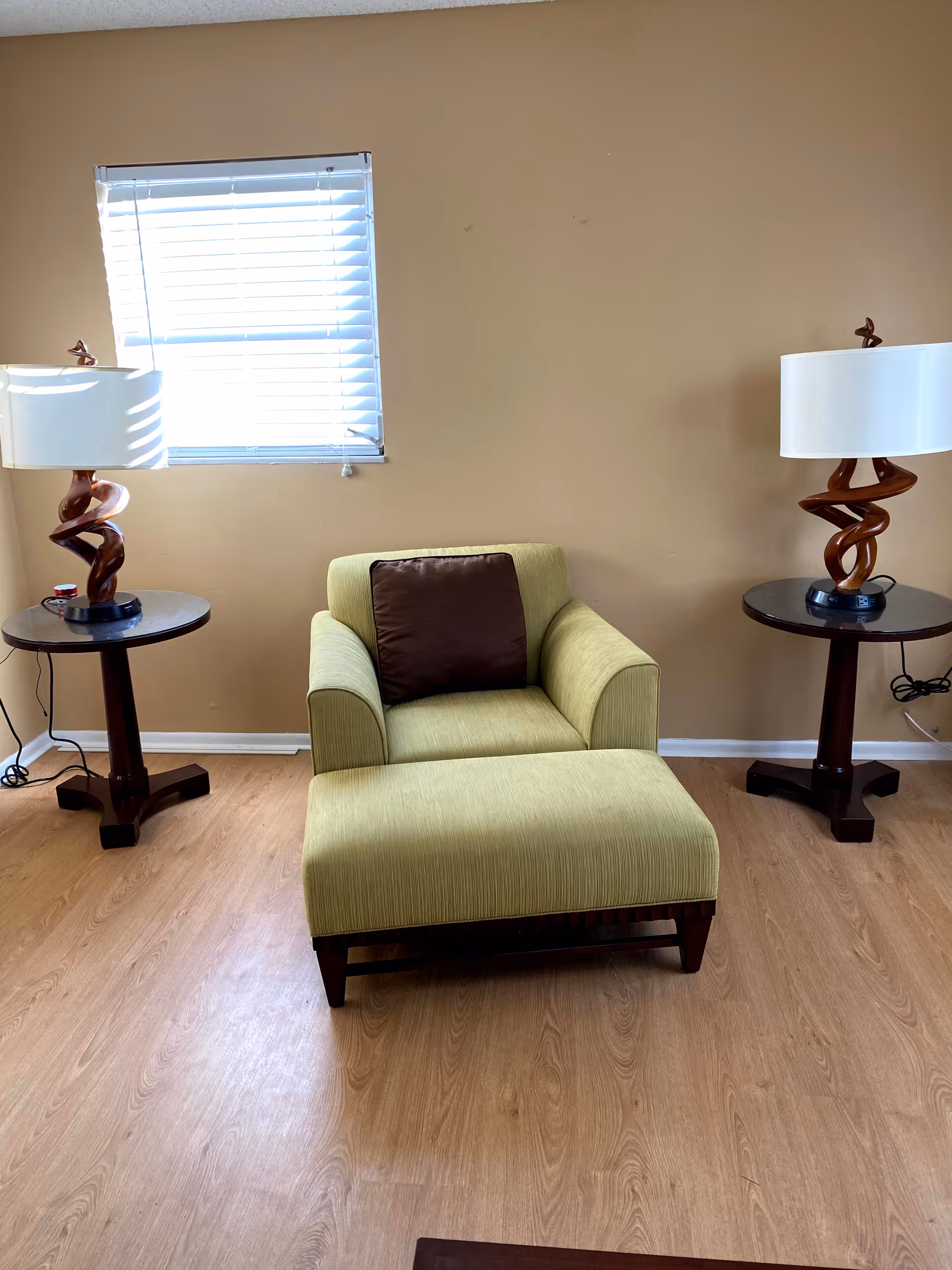 Green armchair with ottoman between two side tables and lamps in a beige living room.