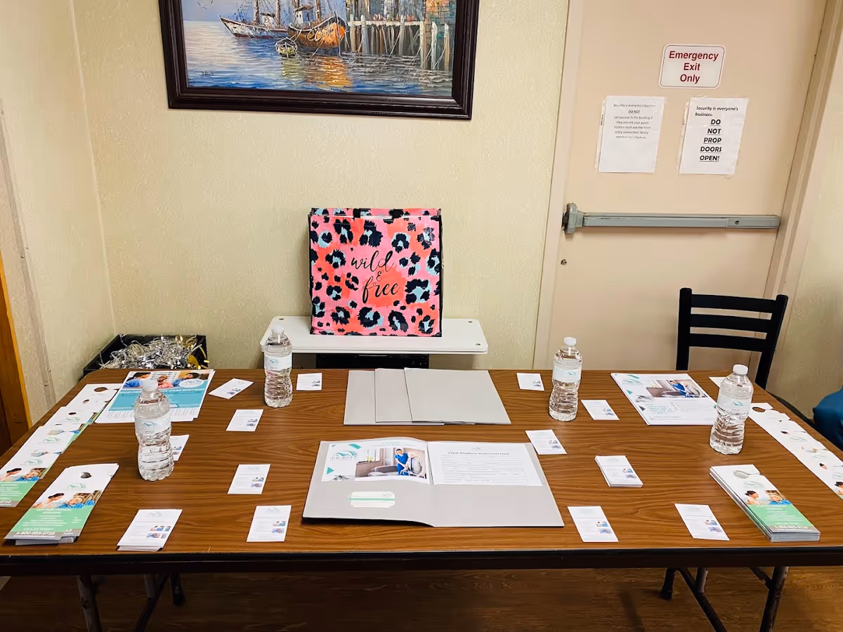 A large wooden table in a room displays brochures, water bottles, and a pink leopard bag.