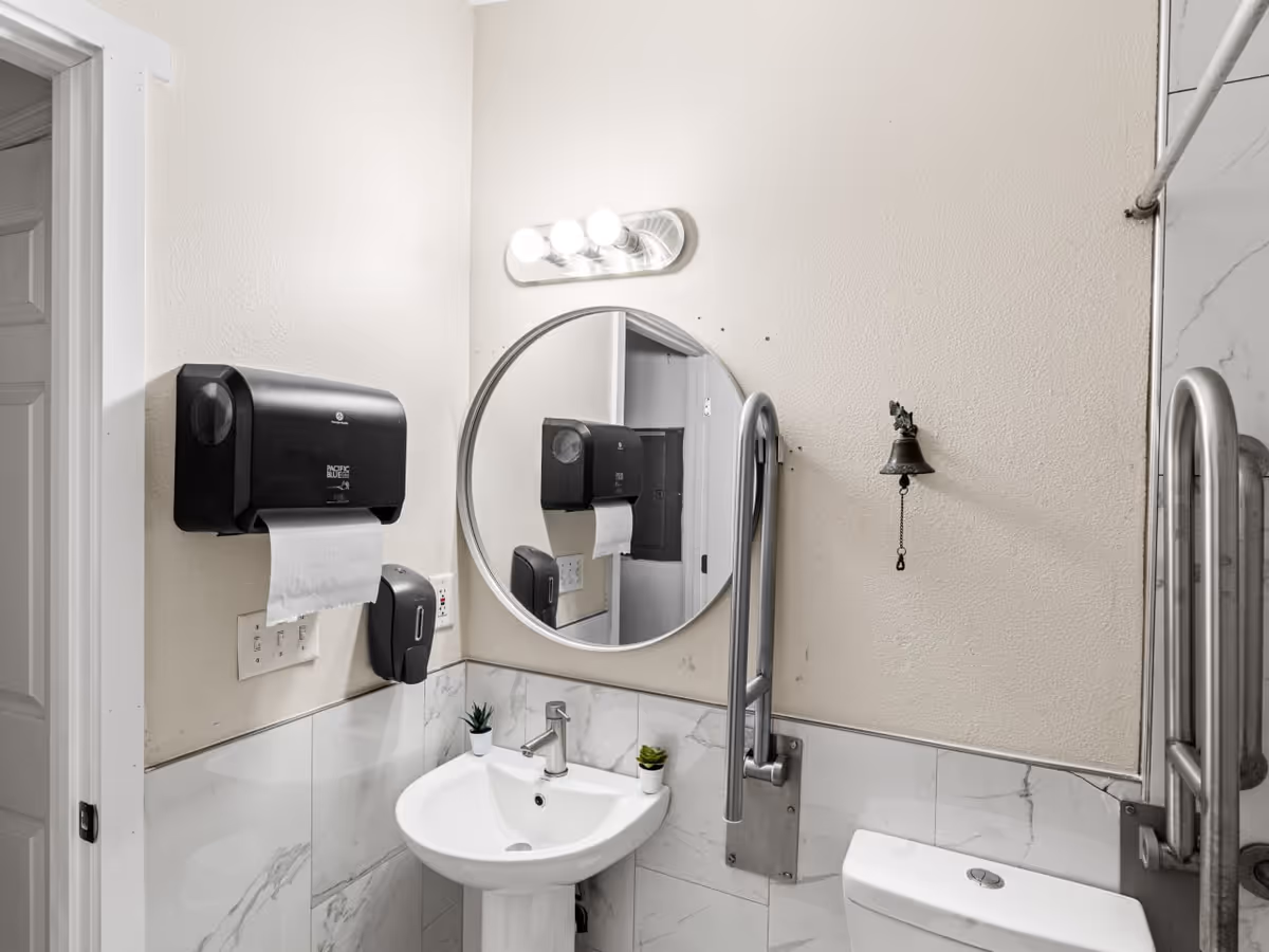 Small bathroom with pedestal sink, round mirror, paper towel dispenser, and grab bar.