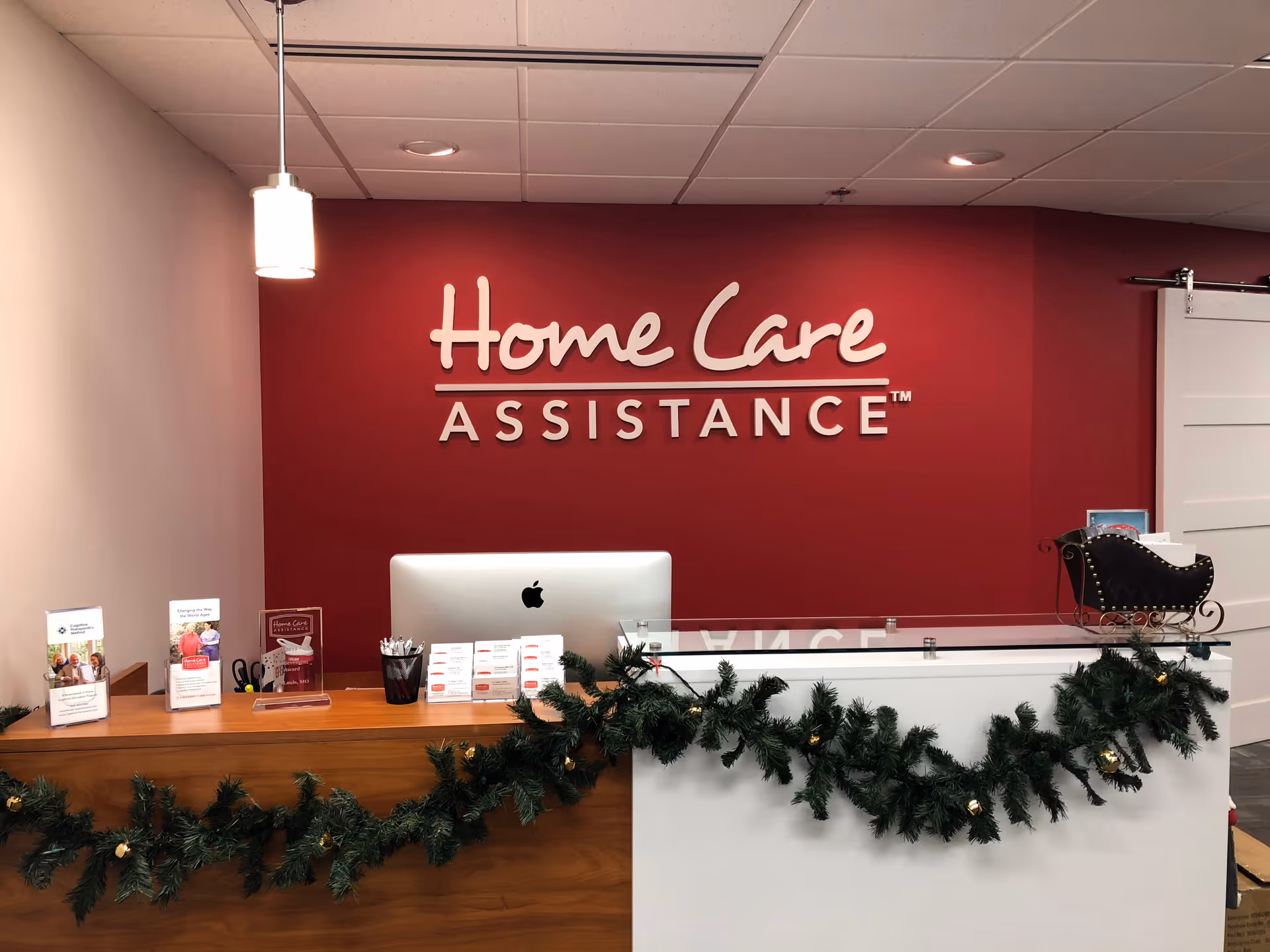 Reception desk in a lobby with a red wall sign reading 'Home Care Assistance' and garland.