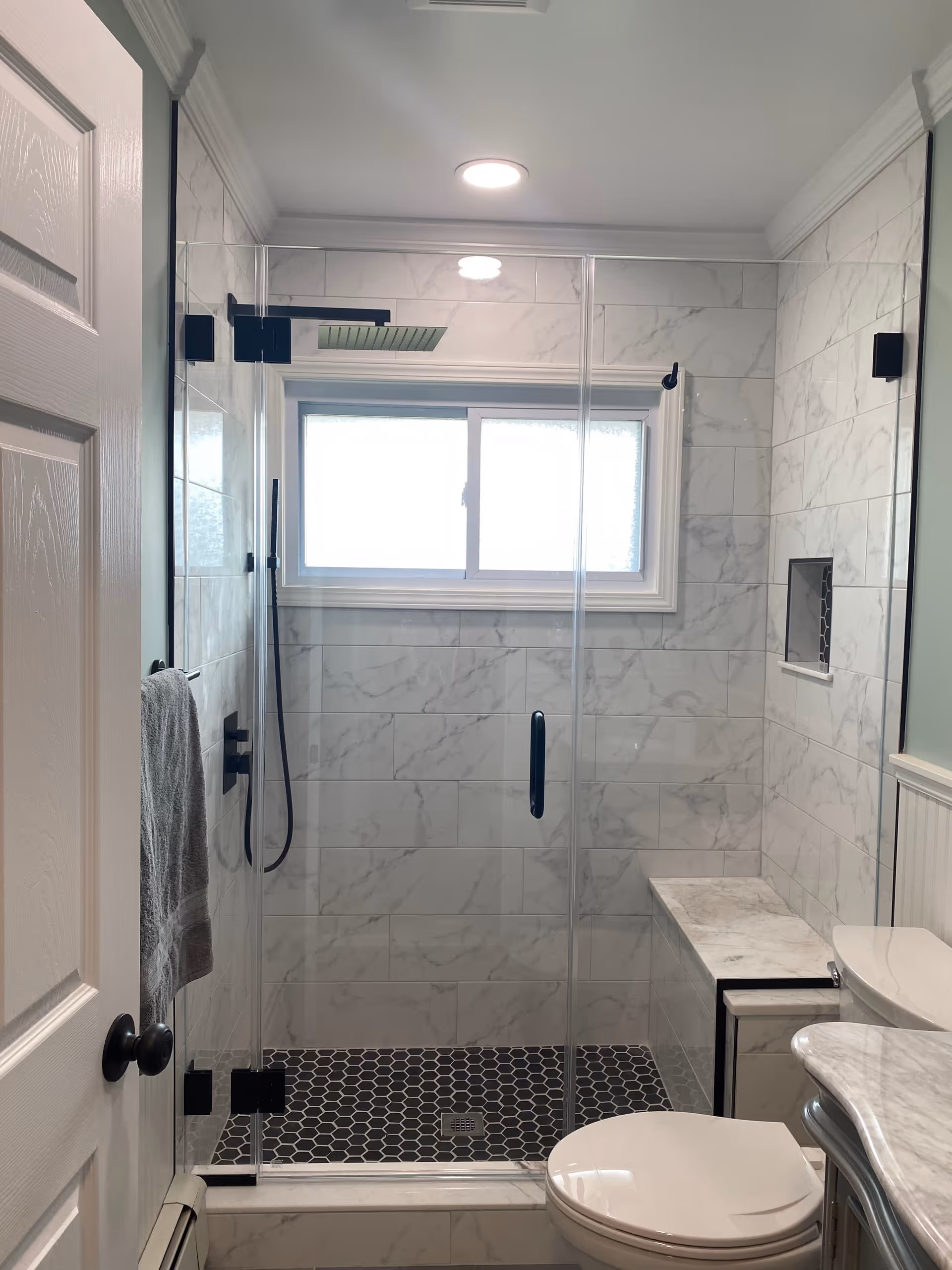 Modern marble-tiled bathroom with glass shower enclosure, bench, toilet, and sink.