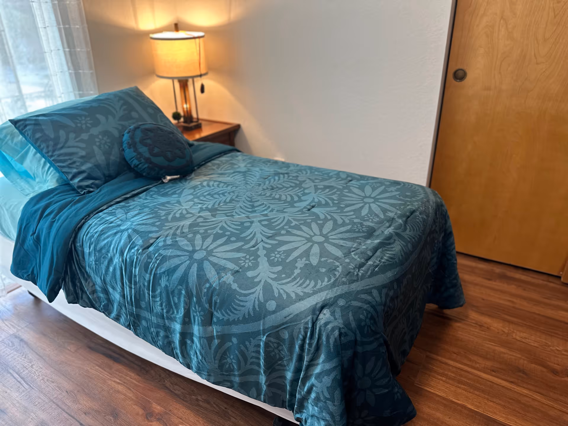 A teal patterned bed with pillows sits in a cozy bedroom beside a wooden door.