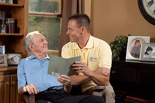 A caregiver and elderly man share a smile in a cozy living room.