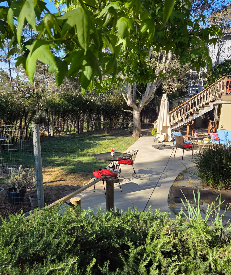 Outdoor patio with tables, chairs, umbrella, and surrounding greenery at a senior living facility.